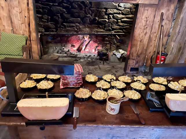 サヴォワ地方のチーズグラタン、奥の暖炉でこんがり仕上げるタルティフレット（Tartiflette）、そして手前の大型チーズ、ラクレット（Raclette）、黒いラクレットグリルはアーム部分の下が熱くなり、下のチーズを表面を溶かす。